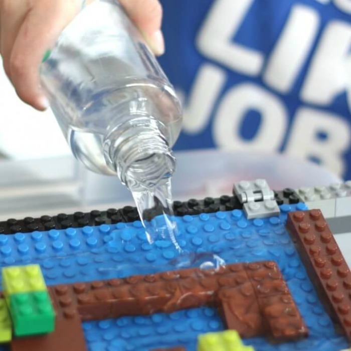 Lego Water Dam Activity for Kids. A hand pouring water to multicolored legos- black, gray, blue, yellow, green and brown